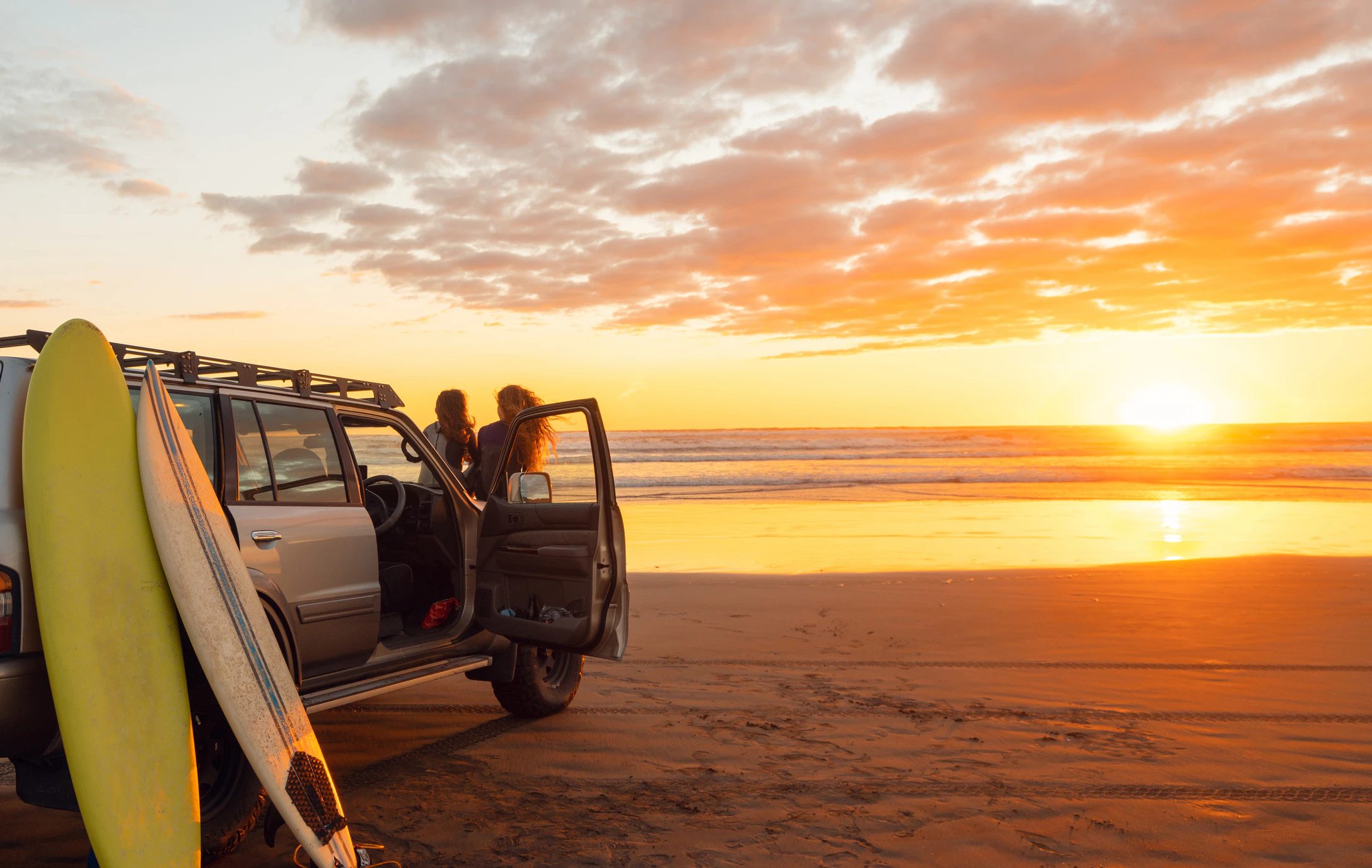 SUV parked on beach at sunset with surfboard and person standing nearby