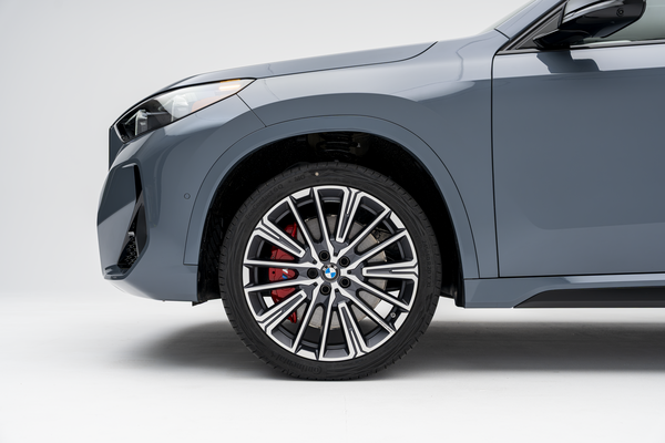 Close-up side view of the front wheel and fender of a grey 2026 BMW X1 xDrive28i, featuring a multi-spoke alloy wheel and red brake caliper.