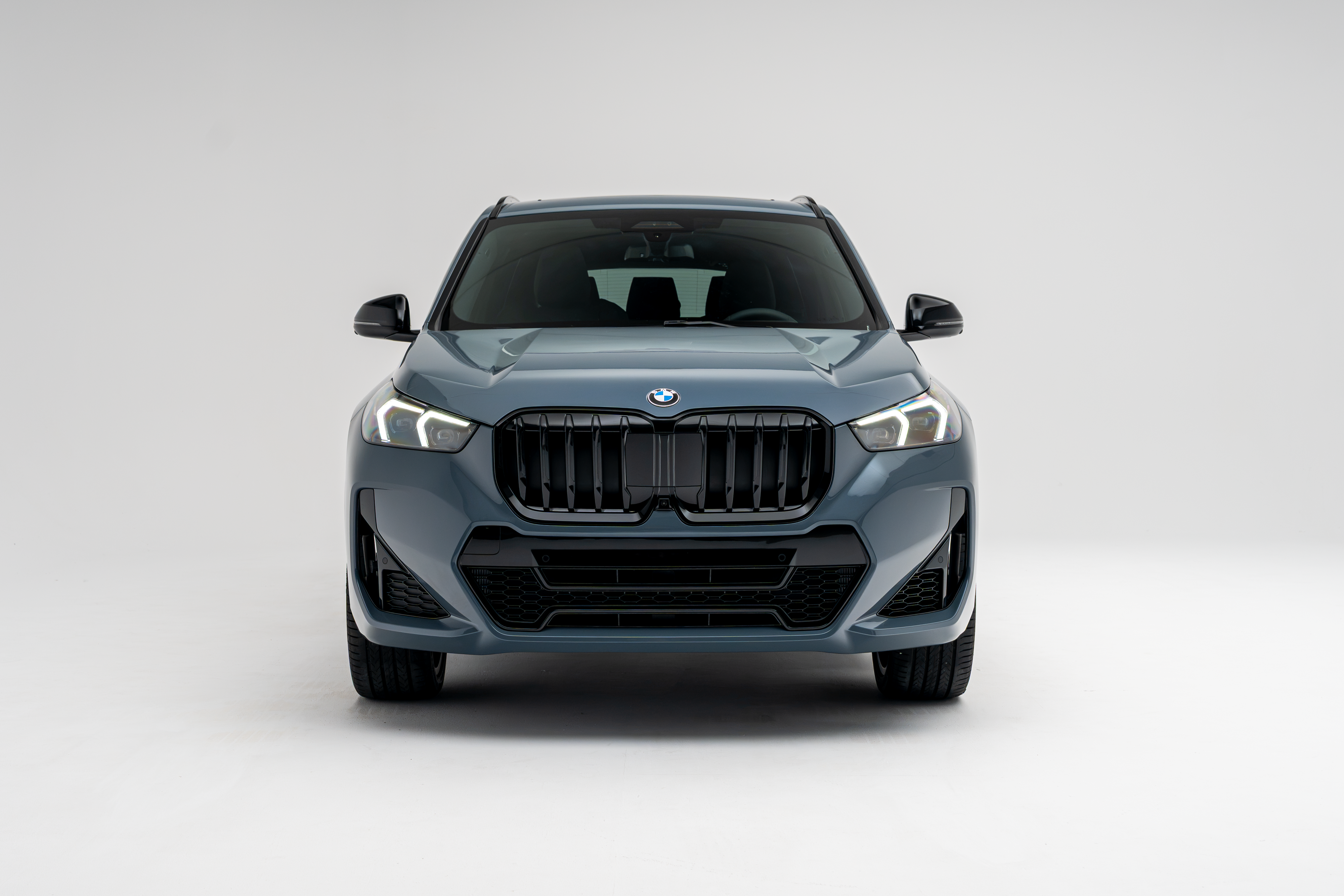 Front view of a grey 2026 BMW X1 xDrive28i SUV with LED headlights illuminated on a white studio background.
