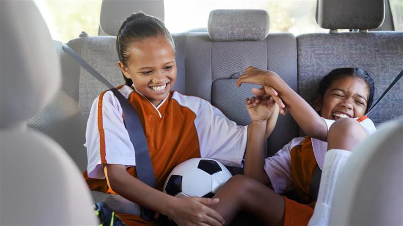Two young girls in sports uniforms playing with a soccer ball in the back seat of a vehicle