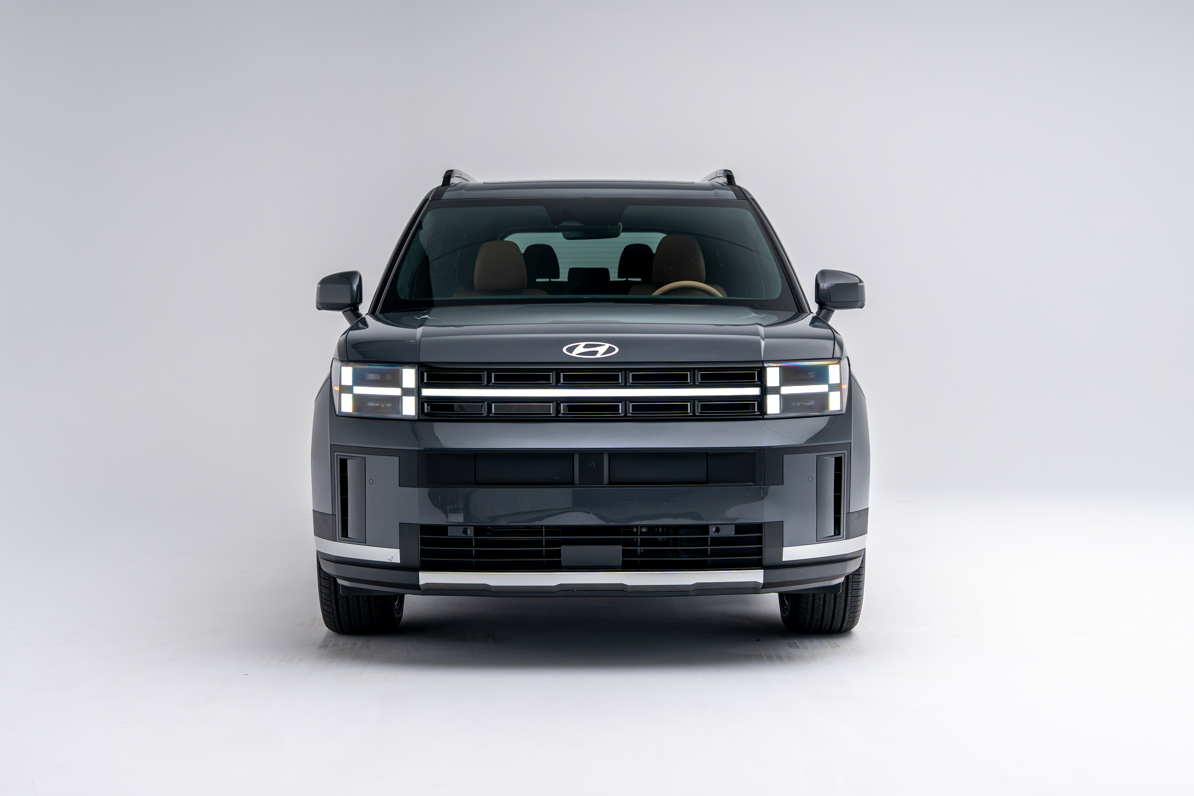 Front view of a grey 2026 Hyundai Santa Fe Hybrid SUV with LED headlights illuminated on a white studio background.