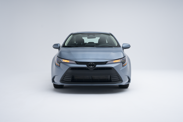 Front view of a light blue 2026 Toyota Corolla with amber lights illuminated on a white studio background.