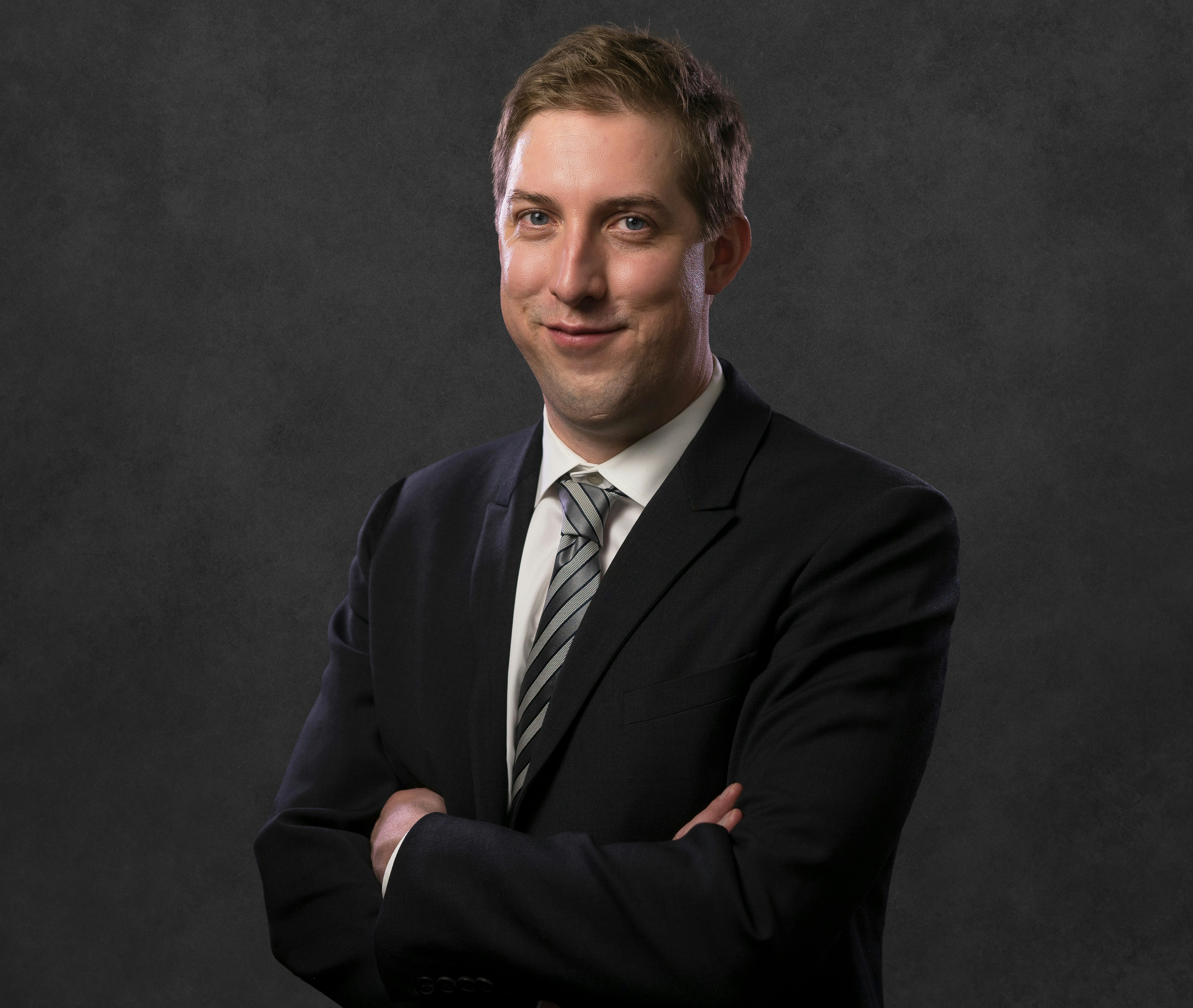 Jared Priestner smiling, arms crossed, wearing a dark suit jacket, white shirt and grey and white tie
