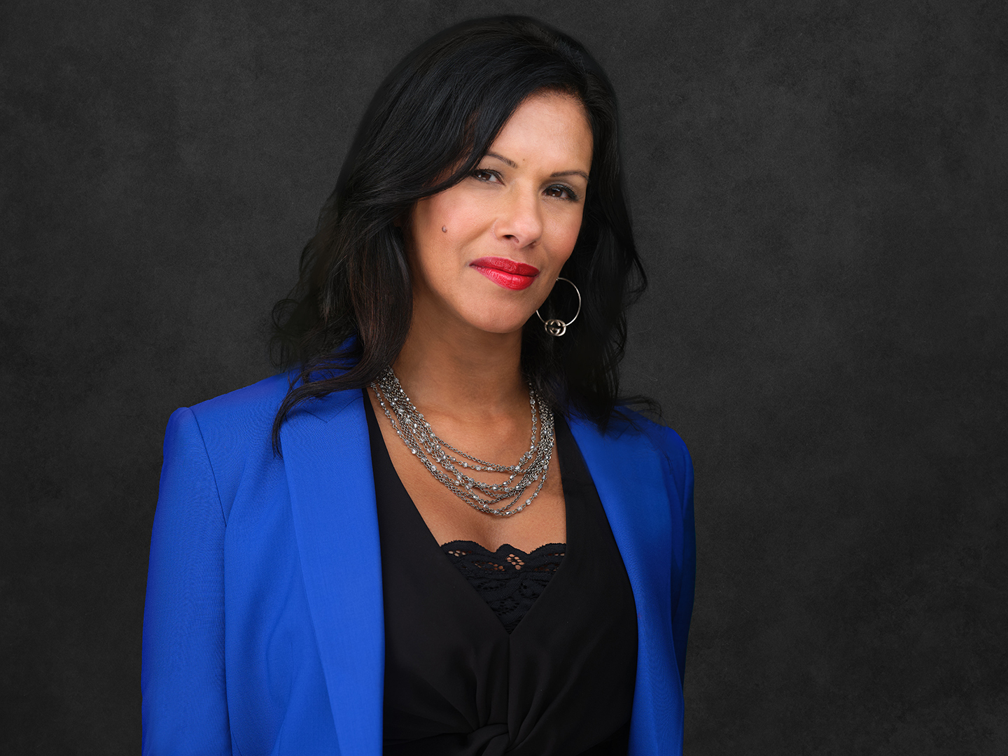 Latha Thomas-Back smiling, wearing red lipstick, earrings, a blue suit jacket and black top