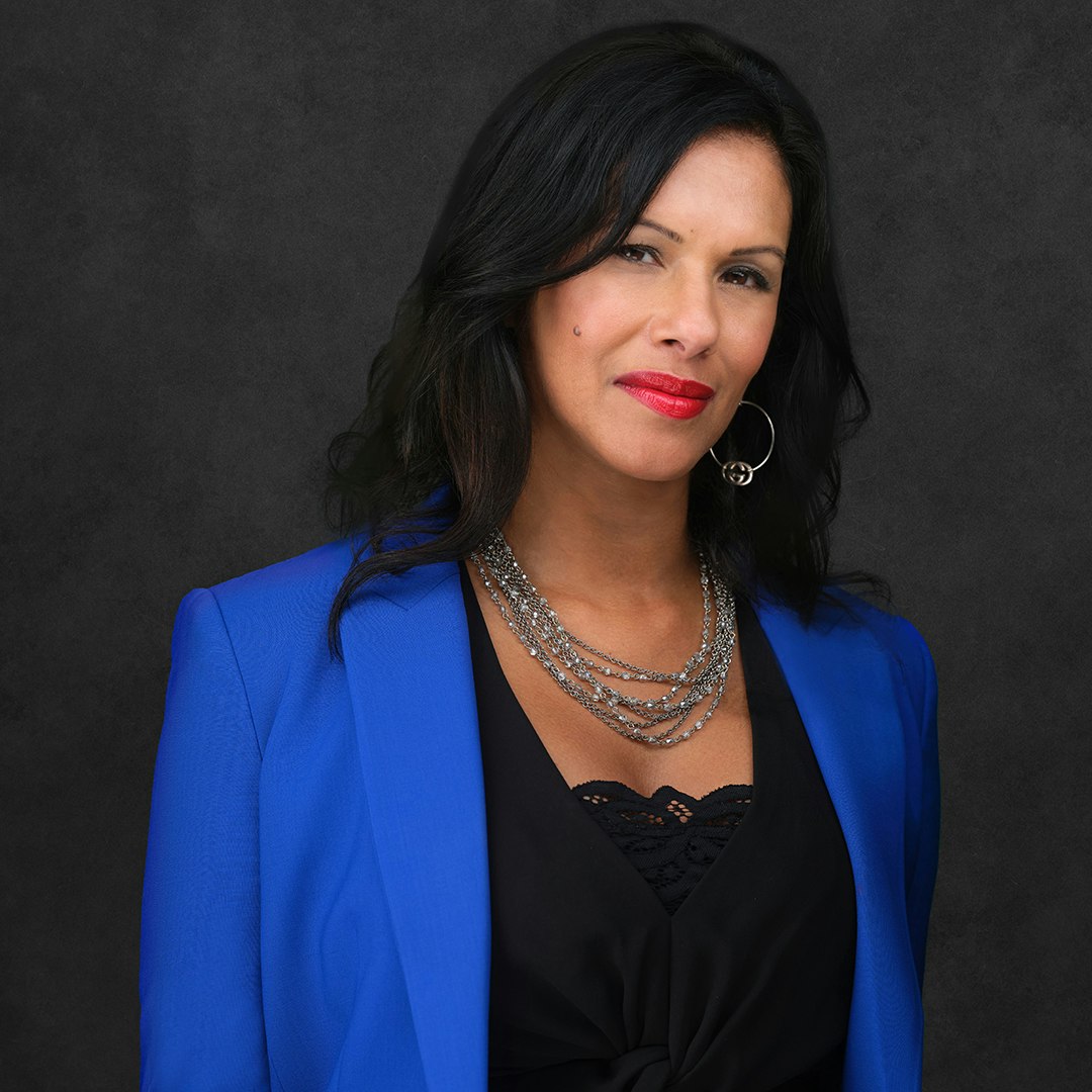 Latha Thomas-Back smiling, wearing red lipstick, earrings, a blue suit jacket and black top