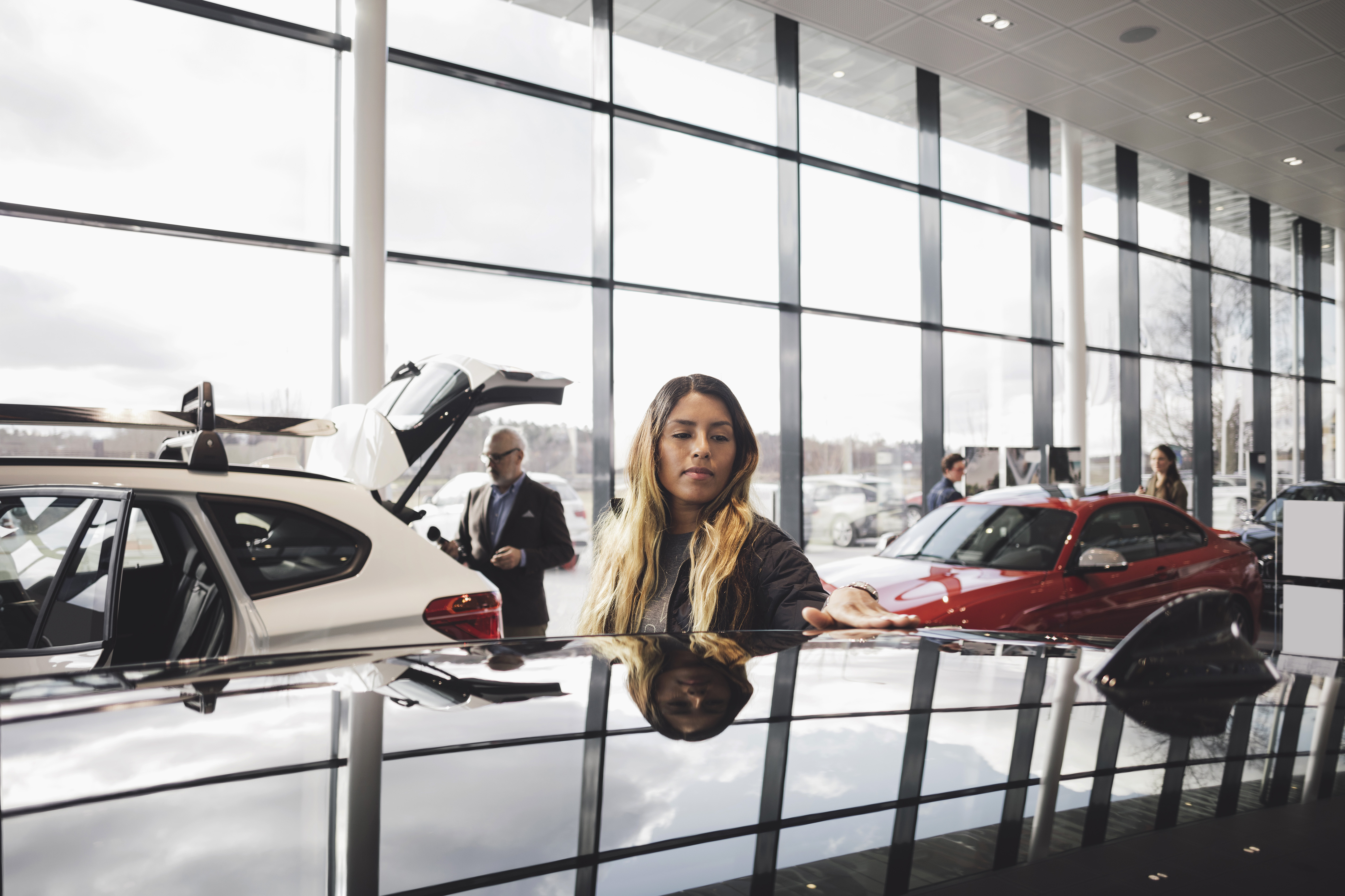 A long-haired woman in a car dealership, looking at a vehicle, with other customers in the background