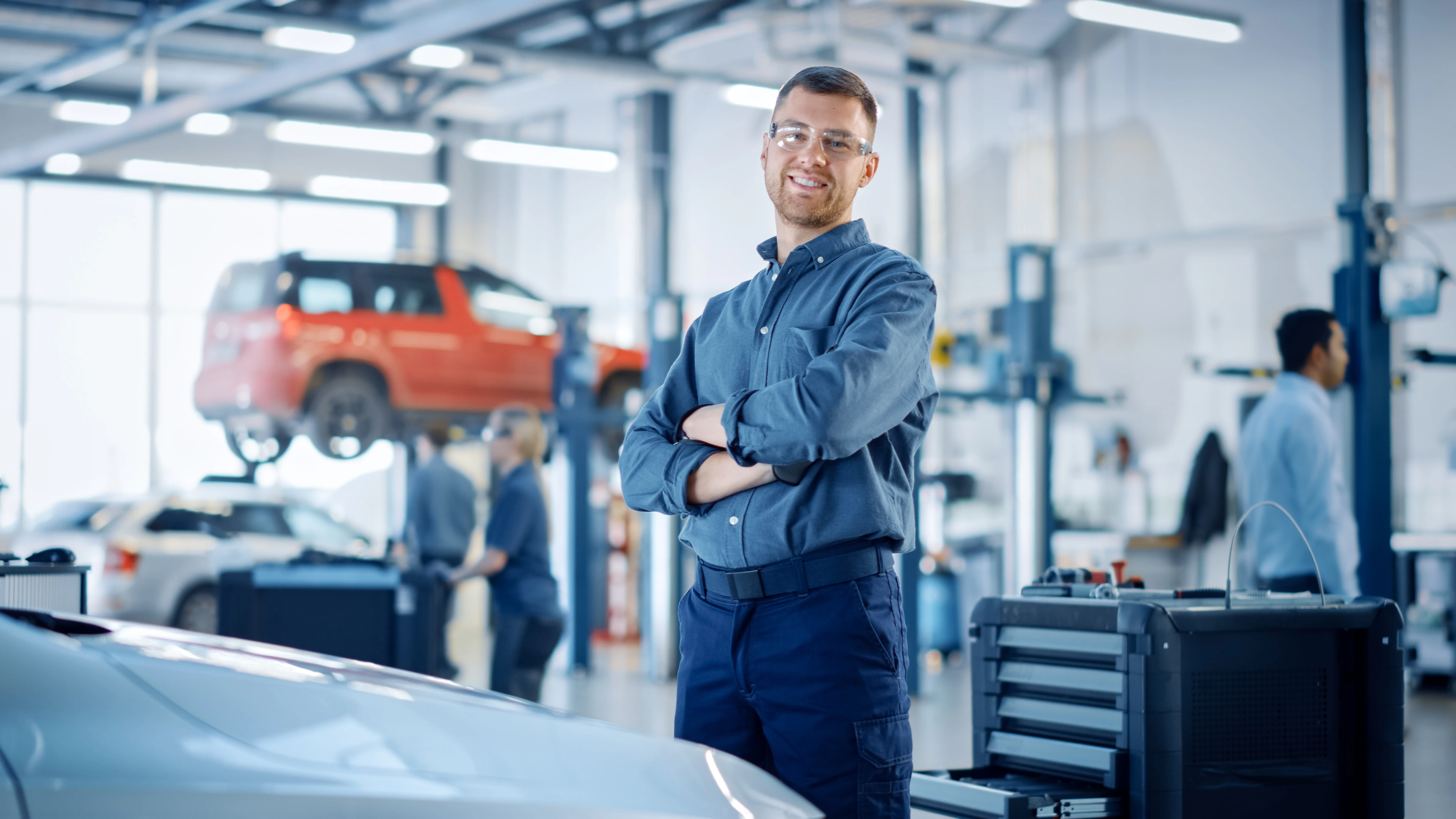 A smiling man in a shirt and trousers stands with arms crossed, facing the camera, with vehicles behind him in a service bay