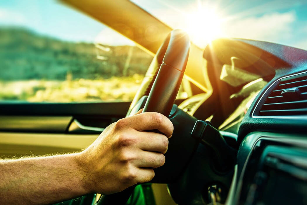 A hand on a vehicle steering wheel, the sun is shining and the driver's window are in the background