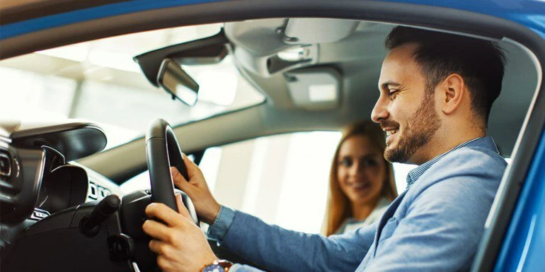 A man in the driver's seat of a parked vehicle, smiling and holding the steering wheel, with a long-haired woman smiling in the passenger seat