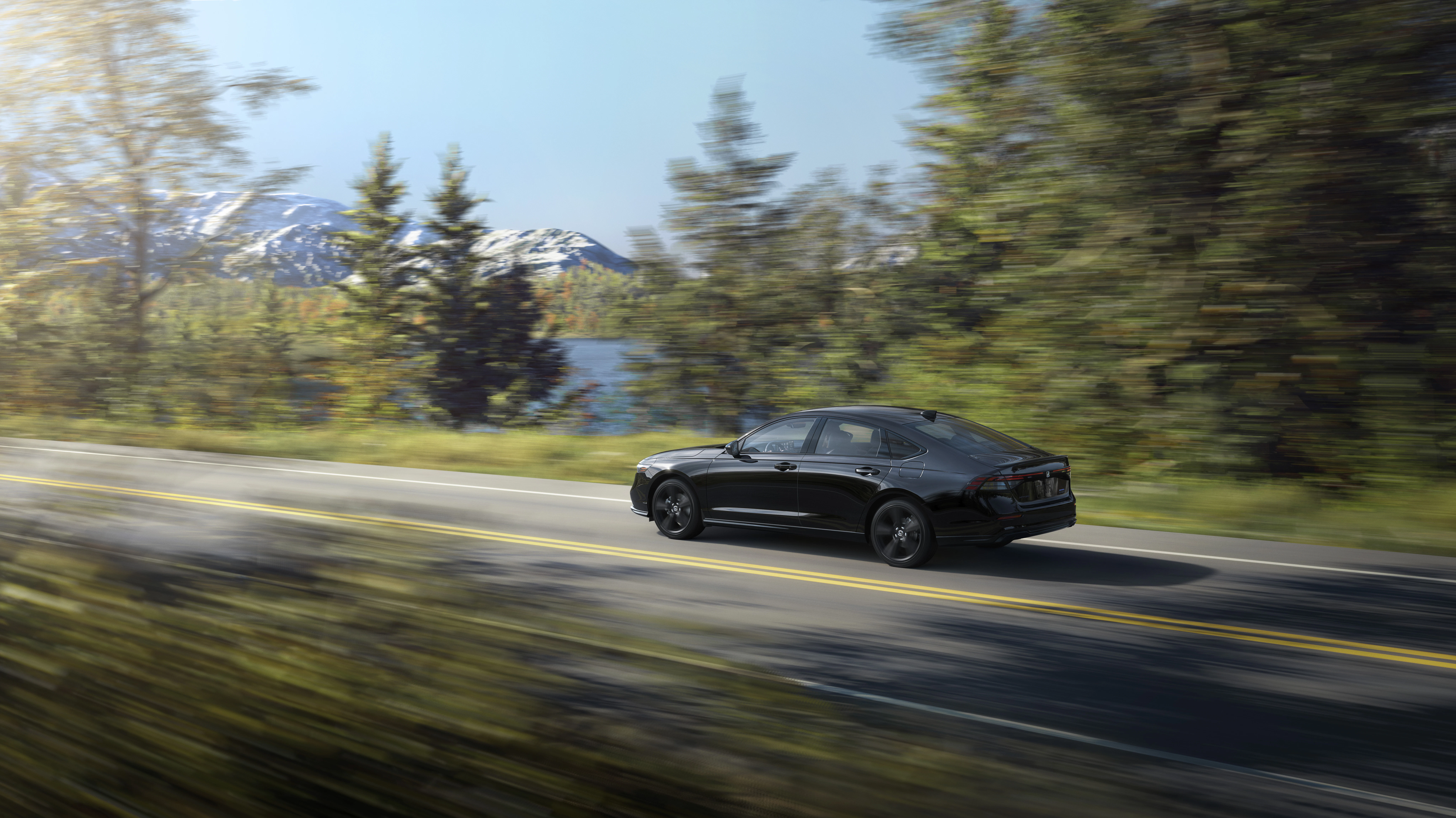 Honda Accord Hybrid Sport cruises along a picturesque mountain roadway, showcasing sleek design and efficient performance against a backdrop of forests, lakes, and snow-capped peaks.