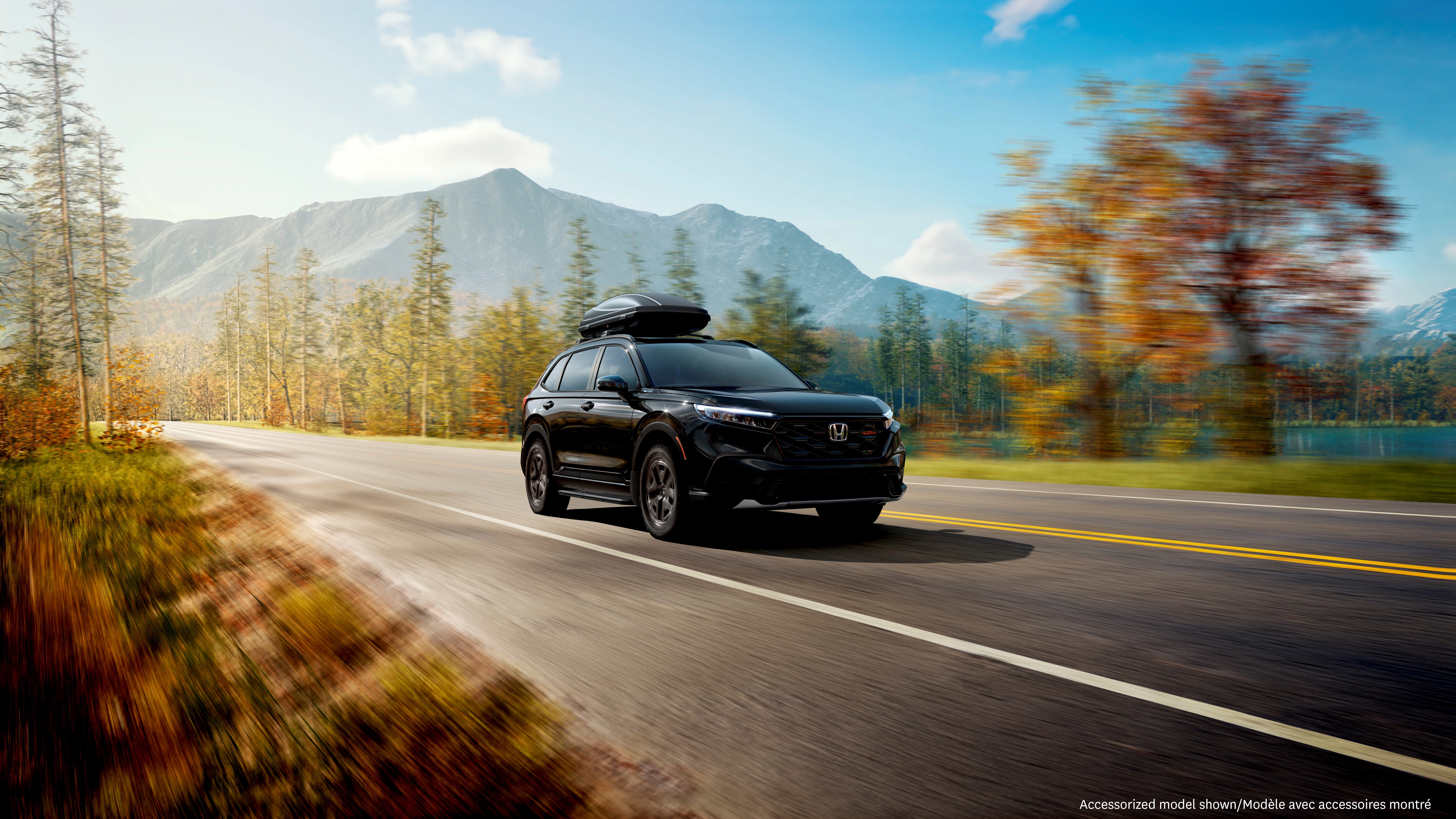 Honda CR-V TrailSport travels along a scenic mountain highway with a rooftop cargo carrier, showcasing adventure-ready versatility amid lakeside views and autumn foliage.