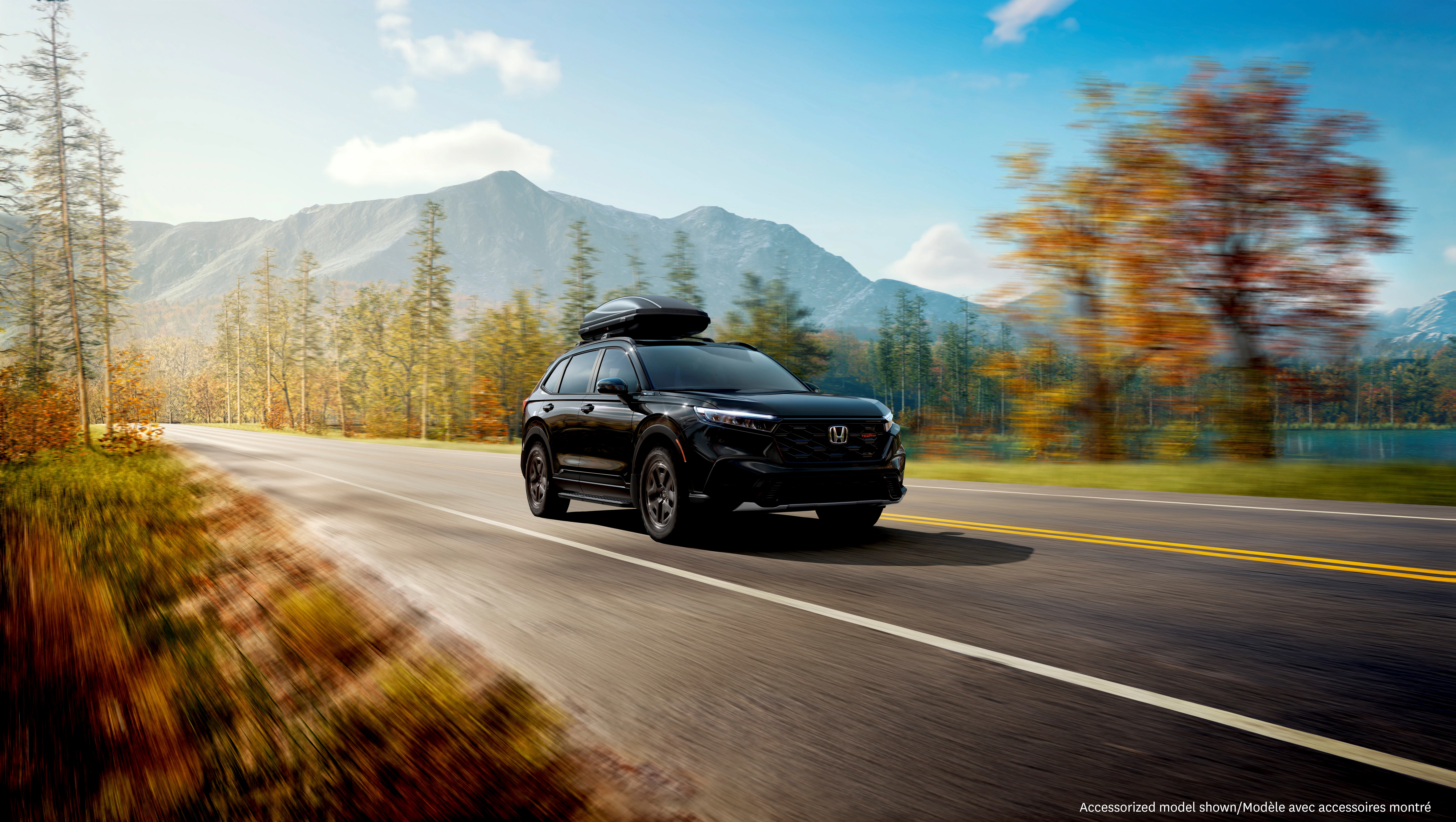 Honda CR-V TrailSport travels along a scenic mountain highway with a rooftop cargo carrier, showcasing adventure-ready versatility amid lakeside views and autumn foliage.