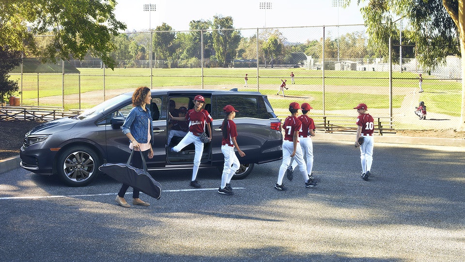A woman walks behind a group of children dressed in baseball gear, getting out of a grey Honda minivan and walking toward a ball diamond.