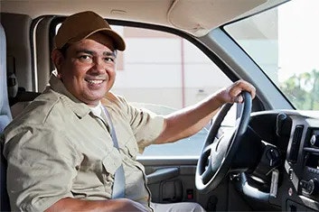 A smiling man in a baseball cap and shirt is in the driver's seat of a parked vehicle, looking at the camera, one hand on the steering wheel