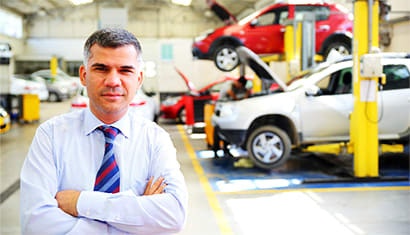 A friendly man in a suit looks at the camera with his arms crossed. Cars are raised up being serviced behind him
