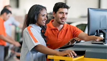 A smiling woman is controlling a computer, and her smiling male colleague is looking at her screen. A vehicle service department is in the background