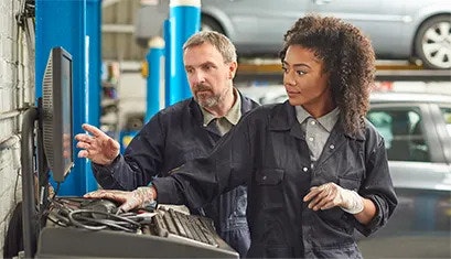 Two workers in overalls in a bodyshop. The female is controlling a computer, and the male is looking at the screen