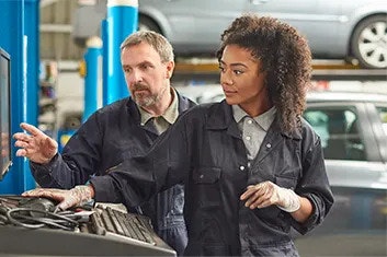 Two workers in overalls in a bodyshop. The female is controlling a computer, and the male is looking at the screen