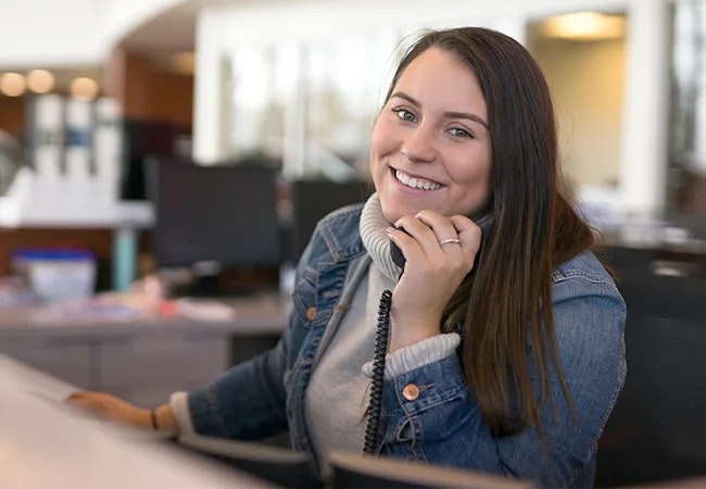 A long-haired woman in a hoodie and denim jacket, sitting in a chair at a reception desk, smiling at the camera