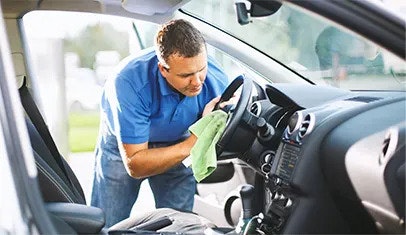 A man in a blue polo shirt and jeans is standing and cleaning the steering wheel of a vehicle with a green cloth