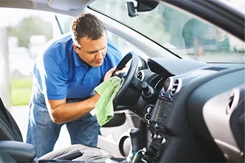 A man in a blue polo shirt and jeans is standing and cleaning the steering wheel of a vehicle with a green cloth