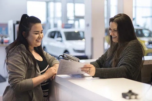 A smiling woman at a counter shows a document to a smiling woman. There are vehicles inside, in the background