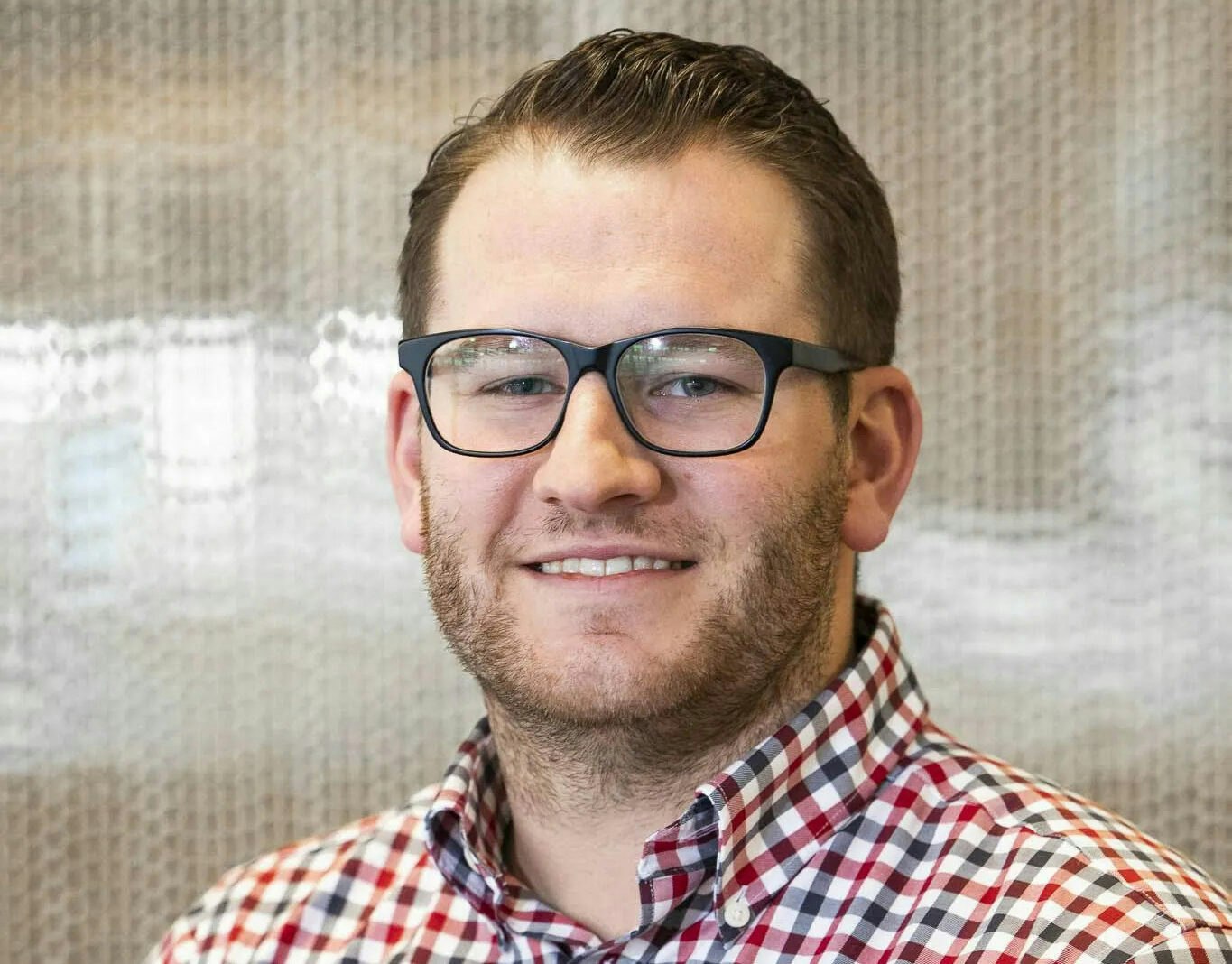 A smiling man with glasses and a beard is wearing a chequered blue, red and white shirt, and looking at the camera