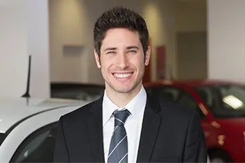 A smiling man in a dark suit, white shirt, and dark striped tie is standing in front of cars, looking at the camera