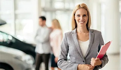 A woman in a suit is holding a red folder and smiling at the camera, two customers are looking at cars behind her