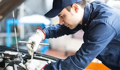A man in blue overalls and a baseball cap is servicing a vehicle under it's hood