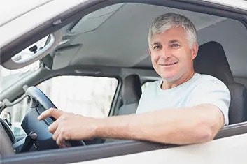 A smiling man is in the driver's seat of a van, holding the steering wheel, looking out the open window at the camera