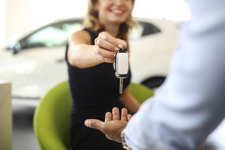 A smiling woman is handing a set of keys to an outstretched hand in front of her. There is a car in the background