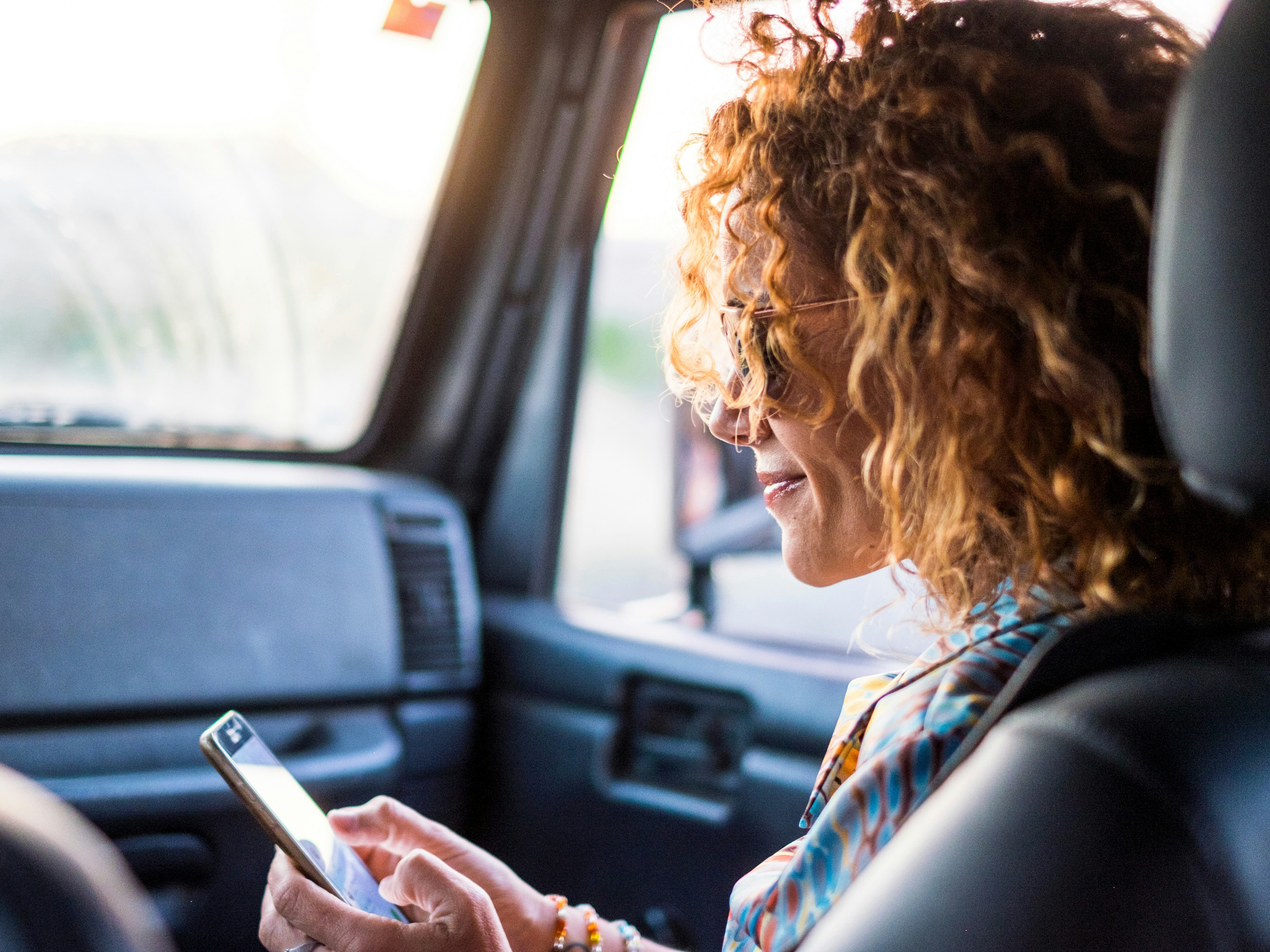 A smiling woman in sunglasses is using her mobile phone in the passenger seat of a vehicle