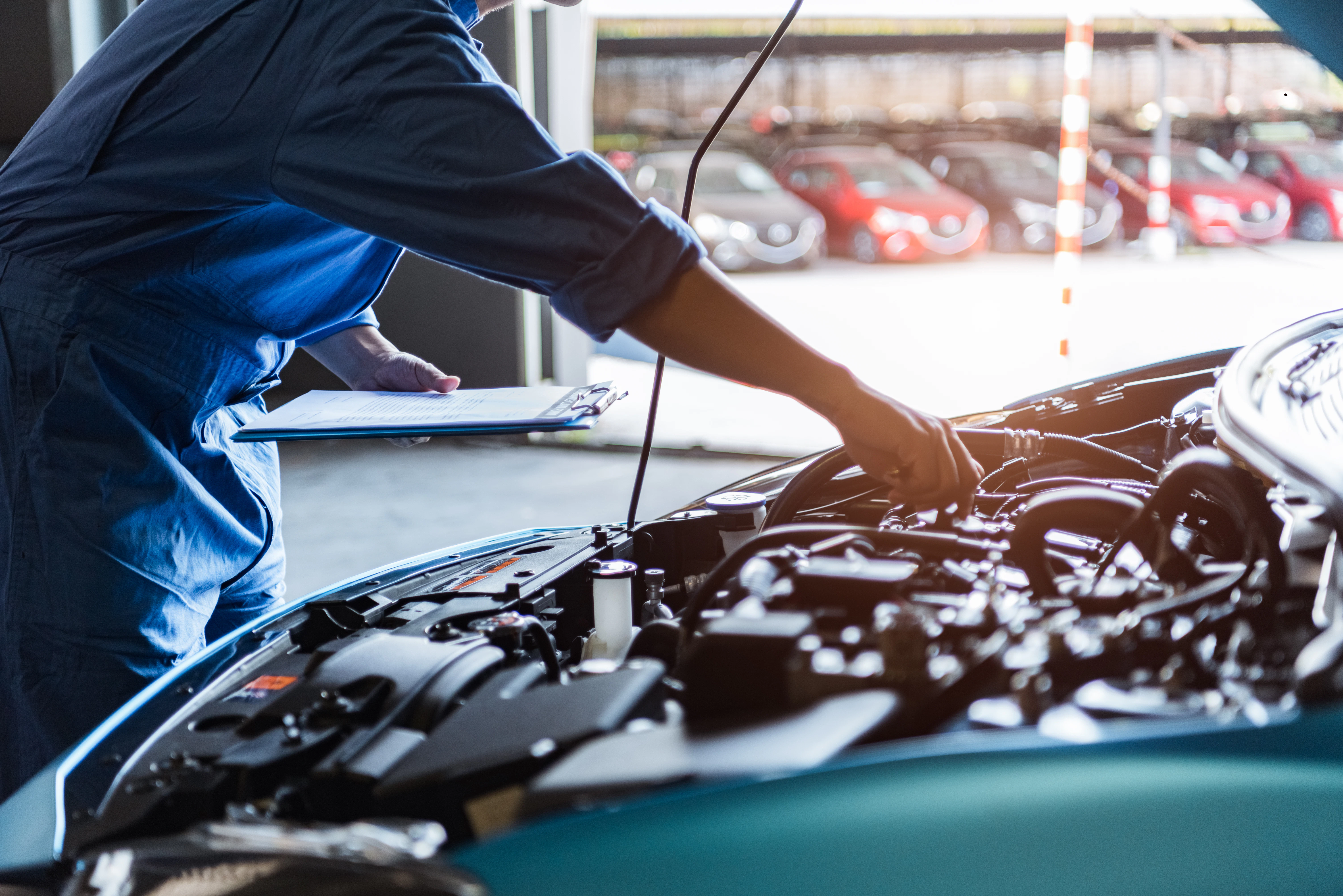 A person in blue overalls is leaning over the open hood of a vehicle, servicing it. Other vehicles are in a service bay in the background