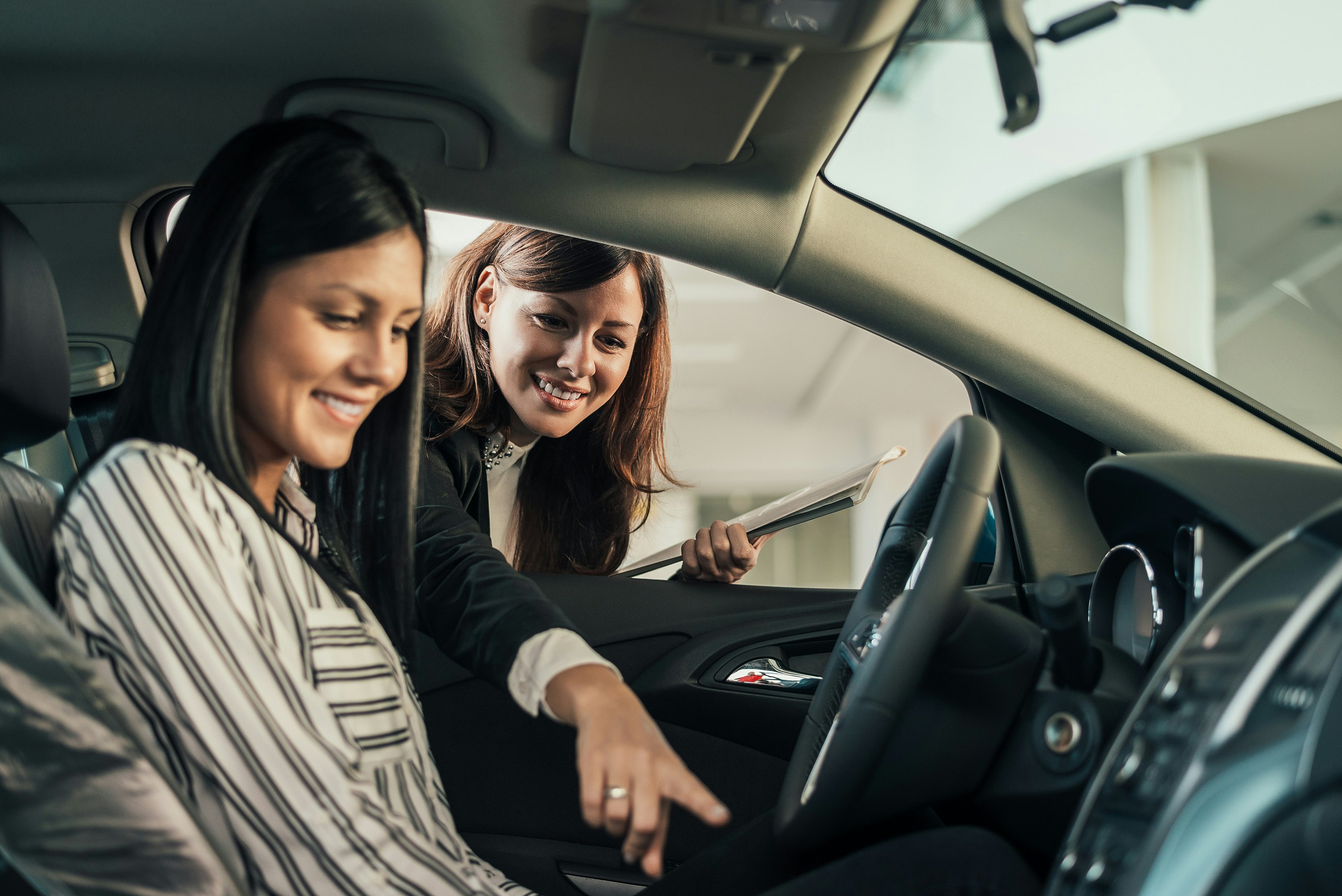 A woman in a striped shirt is in the driver's seat of a vehicle. Another woman outside the vehicle points at the steering wheel