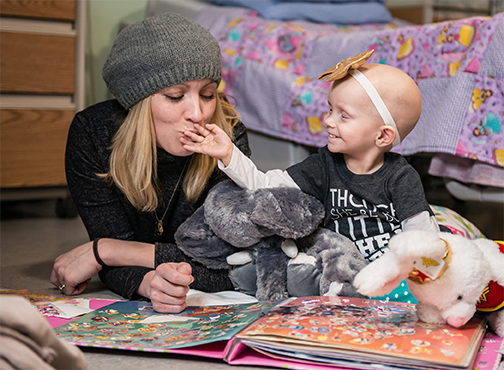 A woman on the left-hand side laying on her stomach, playing with toys, reading a book and kissing the hand of a smiling girl sitting with her on the right