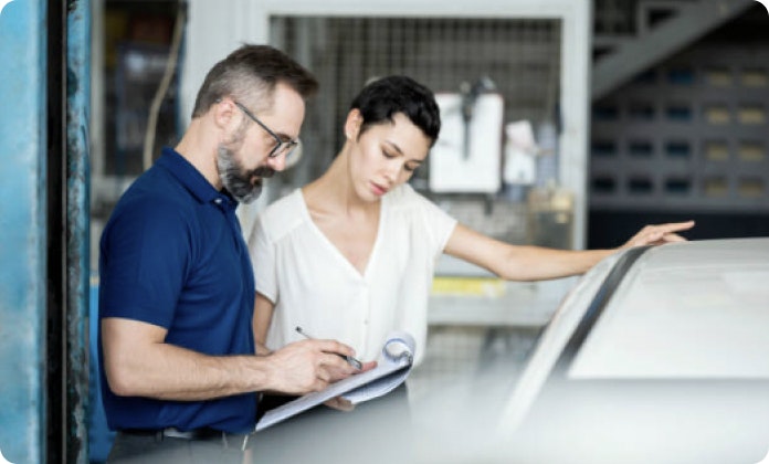 A man is looking at a car and writing on a clipboard. A woman next to him leans on the car and looks at the clipboard
