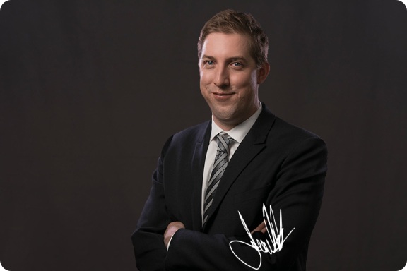 Jared Priestner smiling, arms crossed, wearing a suit. The image is signed by Jared Priestner