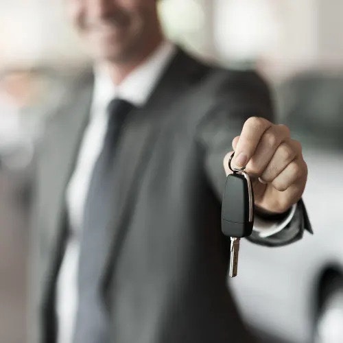 A set of keys is being held out by a smiling man in the background next to a car