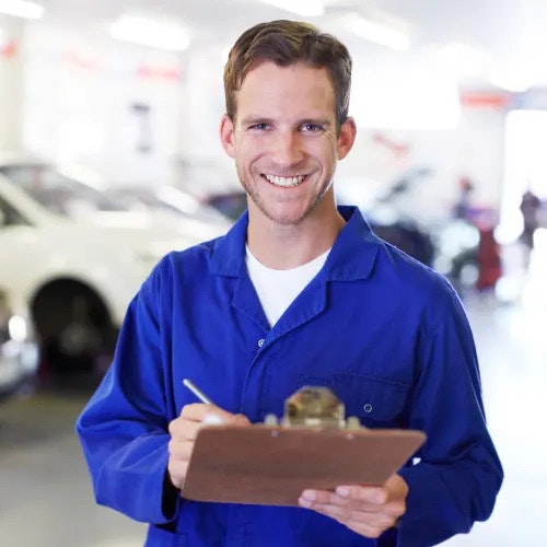A smiling man in a blue work overall is looking at the camera and writing on a clipboard. Vehicles are in a service bay behind him