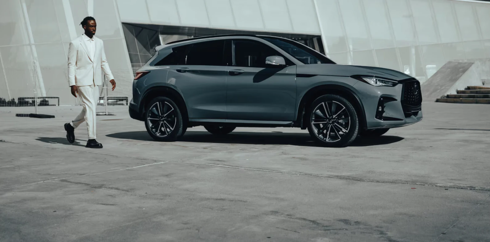 Man in white suit standing beside 2025 Infiniti QX50 SUV in modern architectural setting