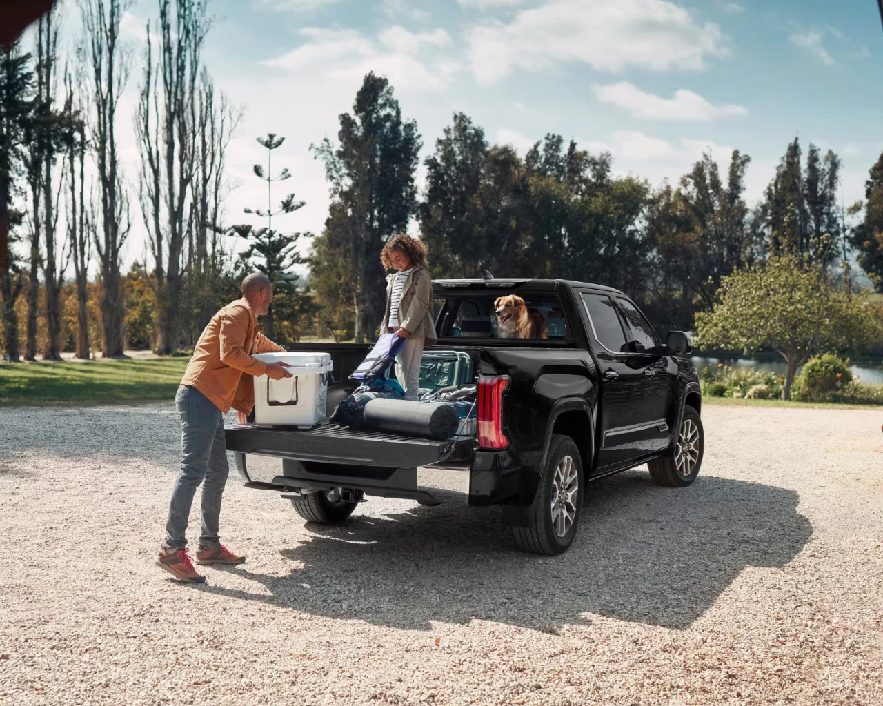 Family loading cooler into black pickup truck bed with dog in cab at outdoor park setting