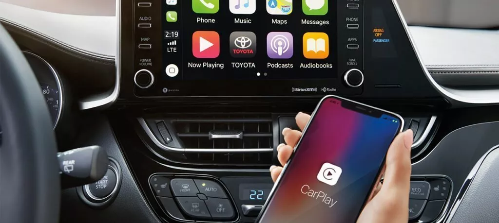 Interior of Toyota C HR with a hand holding an iPhone using CarPlay and the navigation screen in the background