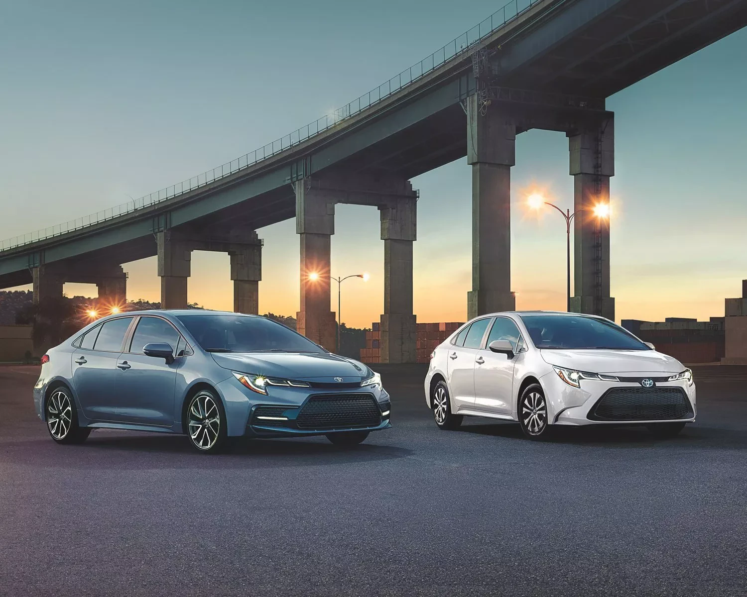 A blue/grey and white Toyota Corolla parked beside each other under a bridge. Sun is going down