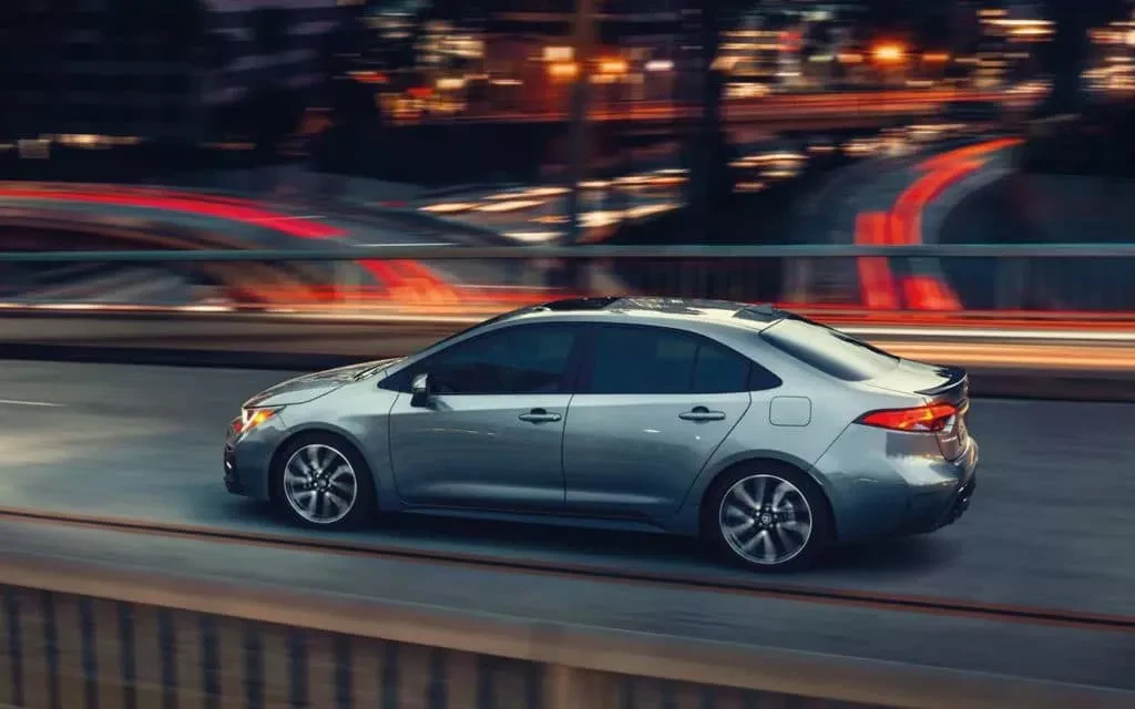 A Toyota Corolla 5 driving down a bridge overlooking a highway at night.