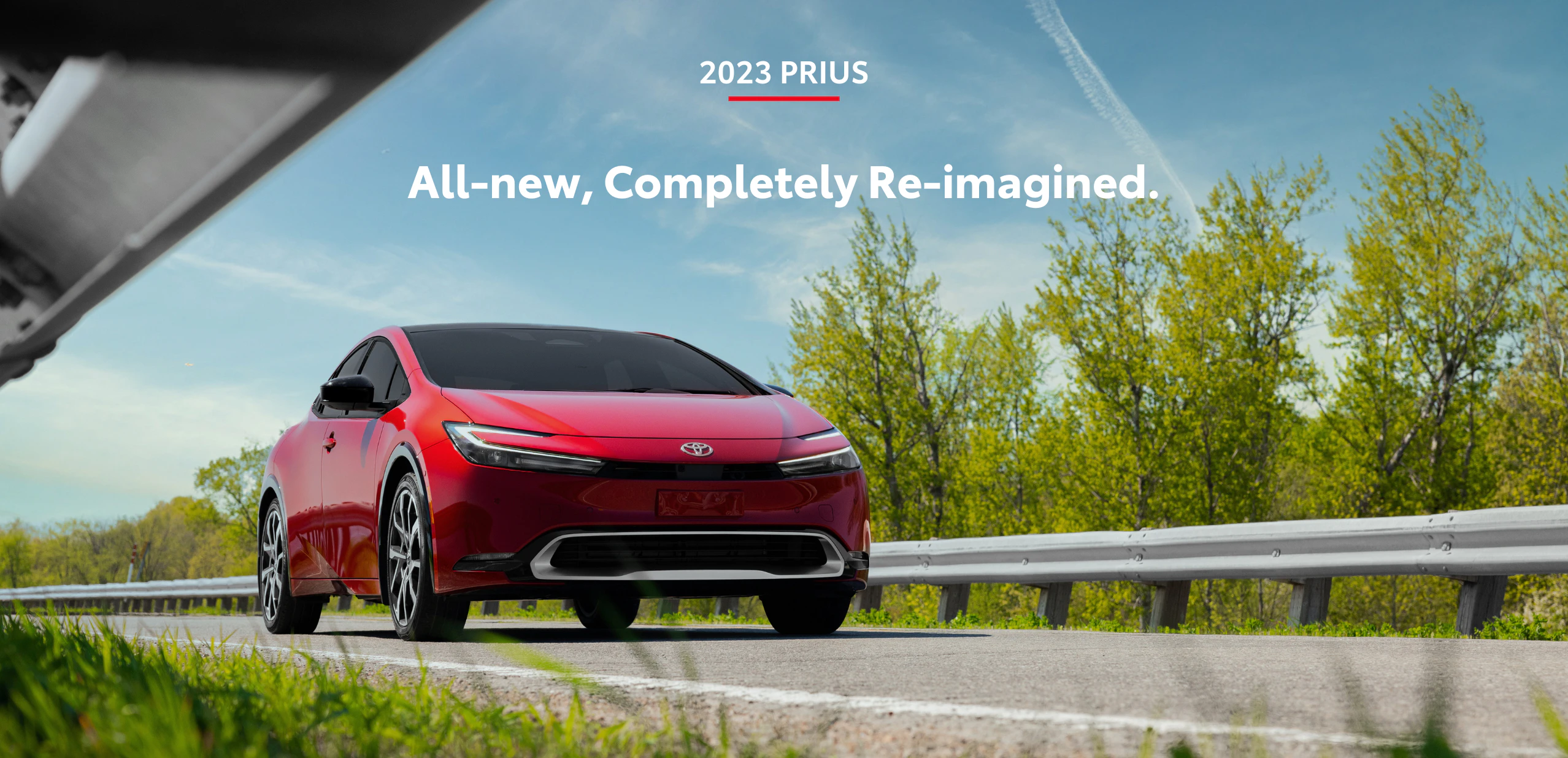 A red Toyota Prius on a road during a bright summer day. Blue sky and trees and grass surrounding the road. Text on image says All new, completely reimagined.