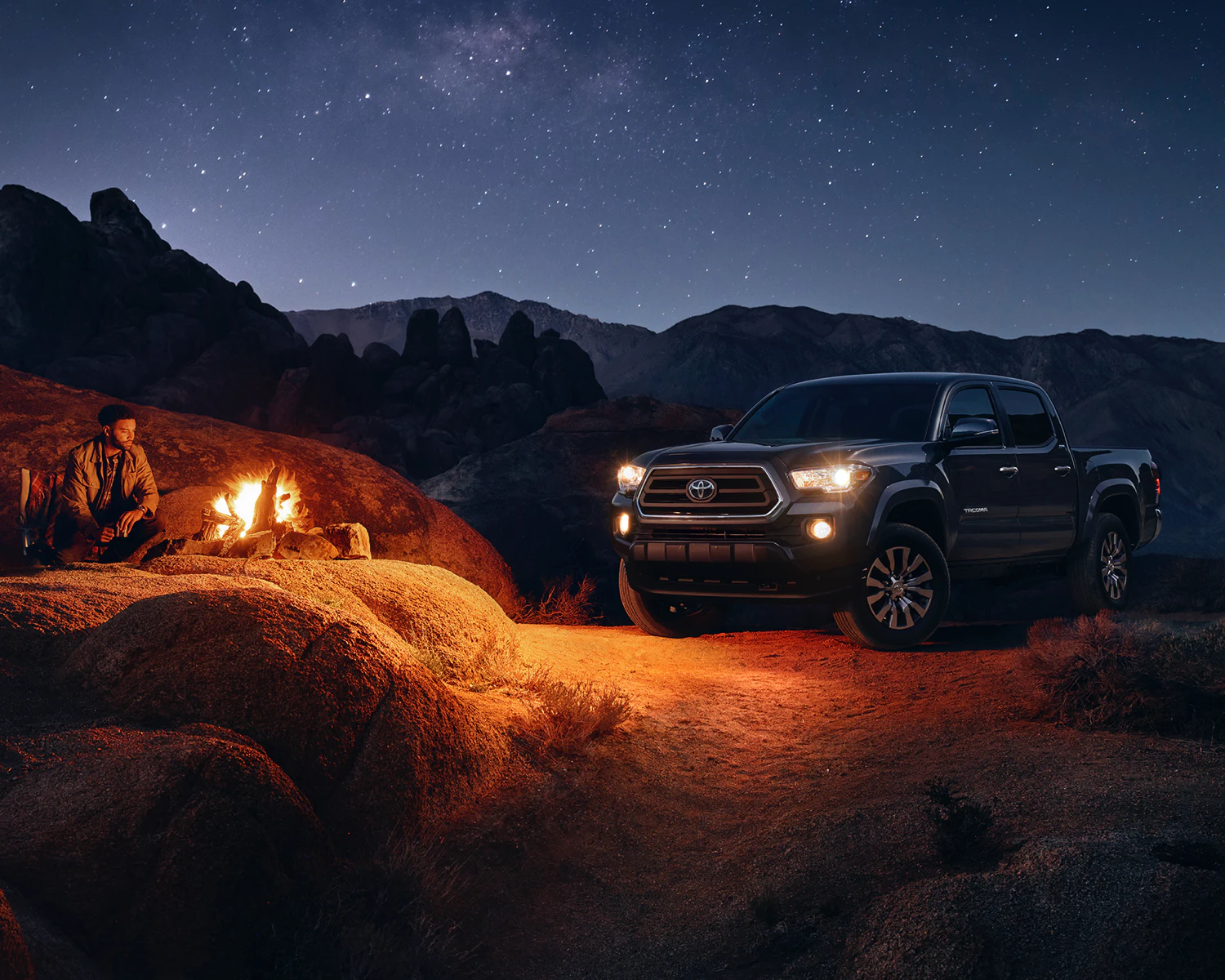 a 2023 Toyota Tacoma parked by a campsite at night where a group of people has a fire going